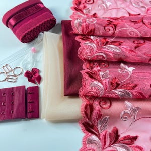 May include: A collection of lingerie-making supplies, including burgundy fabric, floral lace trim, beige mesh, bra hooks, rings, and a small bow. The materials are in shades of burgundy and pink, ideal for crafting custom undergarments.