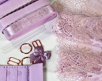 May include: A collection of lavender lingerie making supplies. Includes rolls of ribbon, lace fabric, bra hooks, rings, sliders, and a small bow. The materials are arranged on a white surface.