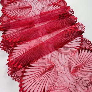 Könnte beinhalten: Ein roter, dekorativer Tischläufer mit einem aufwendigen, gestickten Blumendesign. Der Läufer besteht aus einem transparenten Stoff und hat einen gewellten Rand. Das Design umfasst geschwungene Linien und blattartige Formen.