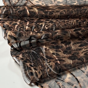 Może przedstawiać: Zbliżenie na przezroczystą tkaninę z wzorem w cętki. Tkanina jest mieszanką brązu i czerni ze złotymi metalicznymi akcentami. Materiał jest złożony, pokazując fakturę i wzór tkaniny.