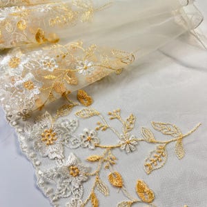 May include: Close-up of ivory fabric with gold and silver floral embroidery. The design features detailed flowers, leaves, and vines, creating a luxurious aesthetic. The fabric is layered, showcasing its texture and depth. Ideal for crafting.