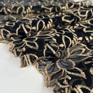 Puede incluir: Primer plano de tela de encaje negro con bordado floral dorado. El delicado patrón crea un efecto texturizado, con el hilo dorado que añade un toque de elegancia. La tela está doblada, mostrando los intrincados detalles.
