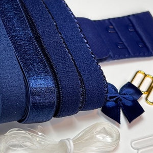 May include: Close-up of navy blue bra-making supplies. The image features navy blue elastic, hook and eye closures, a small bow, gold-colored metal hardware, and clear plastic straps. The materials are arranged on a white surface, suggesting a focus on lingerie or garment construction.