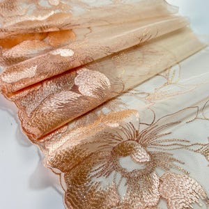 Peut inclure: Gros plan sur un tissu transparent plié avec un motif floral brodé. Le tissu est de couleur pêche clair avec des broderies pêche et dorées. Les bords ont une bordure festonnée.