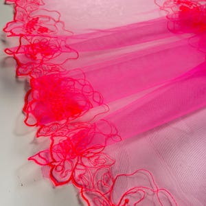 May include: Close-up of bright pink tulle fabric with a floral embroidered edge in a contrasting shade of pink. The sheer material is layered, creating depth and texture. Suitable for crafting or fashion projects.
