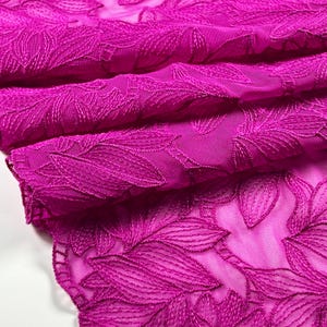 Op de afbeelding: Close-up van een levendige fuchsia stof met een geborduurd bladerpatroon. De stof is opgevouwen en onthult lagen van textuur en detail. Het ingewikkelde ontwerp en de rijke kleur suggereren een hoogwaardige textiel, geschikt voor kleding of decoratieve projecten.
