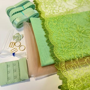 May include: A collection of lingerie-making materials in shades of green and gold. The image shows rolls of ribbon, lace trim, fabric, bra hooks, rings, sliders, and straps. The materials are in shades of green, gold, and beige.