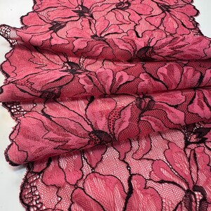 Op de afbeelding: Close-up van een gevouwen stuk roze kanten stof met een bloemenpatroon. Het kant heeft grote, gestileerde bloemen met zwarte contouren. De stof is gelaagd en toont de ingewikkelde details van het kantontwerp.