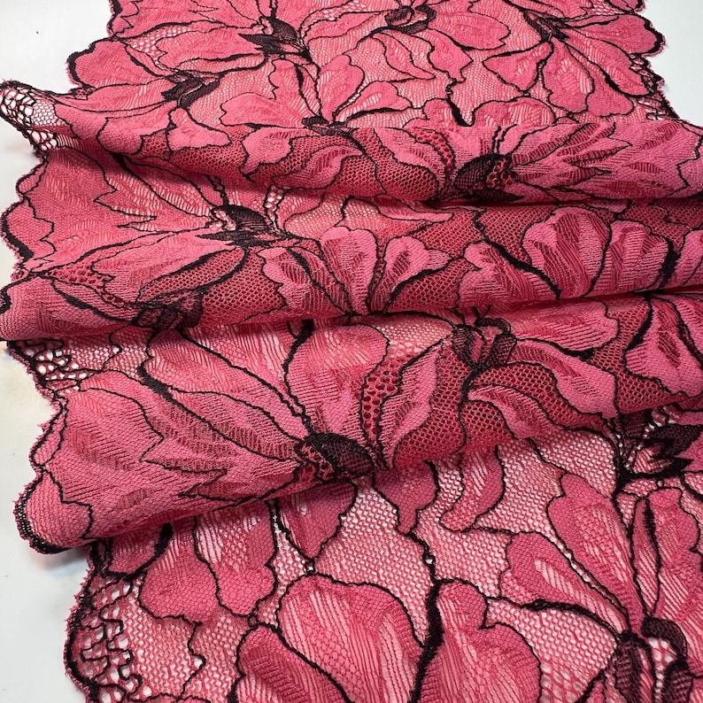 Op de afbeelding: Close-up van een gevouwen stuk roze kanten stof met een bloemenpatroon. Het kant heeft grote, gestileerde bloemen met zwarte contouren. De stof is gelaagd en toont de ingewikkelde details van het kantontwerp.
