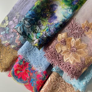 Può includere: Una collezione di campioni di tessuto sovrapposti con intricati ricami floreali su rete trasparente. I tessuti mostrano una varietà di colori tra cui lavanda, blu vibrante, rosa neon, oro e pesca, con bordi smerlati e cuciture dettagliate.