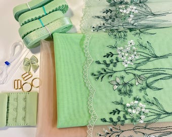 May include: A collection of sewing and lingerie-making supplies in shades of green and beige. Includes rolls of ribbon, fabric, lace with floral embroidery, hooks, eye closures, and metal hardware.