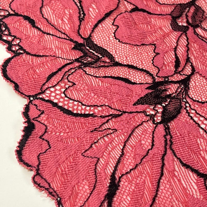 Op de afbeelding: Close-up van roze kanten stof met een bloemenmotief. Het kant heeft een gedetailleerd patroon met zwarte contouren, wat contrasteert met de roze achtergrond. De randen zijn geschulpt.