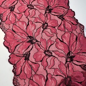 Op de afbeelding: Close-up van een bloemenkanten stof in een dieproze kleur met zwarte contouren. Het kant heeft een herhalend patroon van grote, gestileerde bloemen. De randen van de stof zijn geschulpt, wat een decoratief tintje geeft. De stof wordt waarschijnlijk gebruikt voor kleding of woondecoratie.