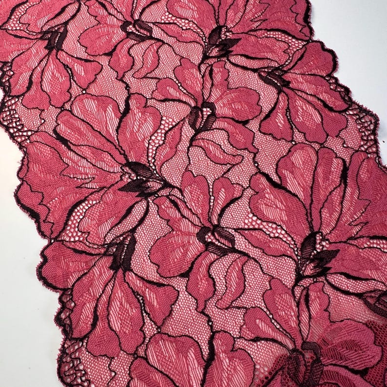 Op de afbeelding: Close-up van een bloemenkanten stof in een dieproze kleur met zwarte contouren. Het kant heeft een herhalend patroon van grote, gestileerde bloemen. De randen van de stof zijn geschulpt, wat een decoratief tintje geeft. De stof wordt waarschijnlijk gebruikt voor kleding of woondecoratie.