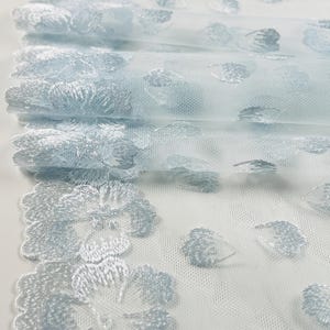 May include: Close-up of a light blue tulle fabric with embroidered floral patterns. The fabric is folded, revealing layers of delicate material. The floral embroidery adds texture and detail to the airy, semi-transparent fabric.