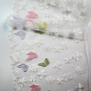 Peut inclure: Dentelle blanche ornée de broderies de papillons et de fleurs roses, vertes et lavandes. La dentelle a un bord festonné avec de petites perles jaunes. Ce tissu délicat est idéal pour divers projets créatifs.