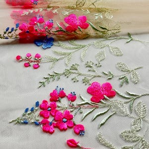 May include: Close-up of floral embroidered fabric. The design features vibrant pink flowers, small blue accents, and green leaves on a beige mesh background. The embroidery is detailed and delicate, showcasing a botanical theme.