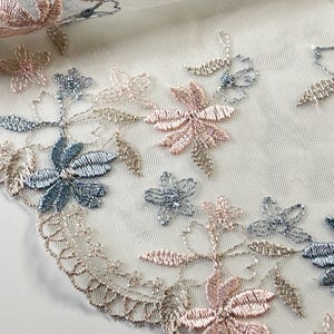 Può includere: Primo piano di delicato pizzo floreale. Il pizzo presenta fiori ricamati nei toni del rosa, blu e oro. I fiori sono disposti in un motivo ripetuto su uno sfondo bianco trasparente. Il ricamo ha una lucentezza metallica.