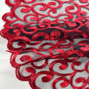 May include: Close-up of red embroidered lace fabric with a swirling, ornate pattern. The design features a repeating motif of stylized curls and curves against a black mesh background. The fabric is layered, creating depth and texture.