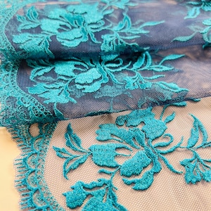 Puede incluir: Primer plano de tela de encaje floral turquesa. El encaje presenta flores y hojas bordadas con un borde festoneado. La tela está en capas, mostrando una malla azul más oscura debajo del bordado turquesa.