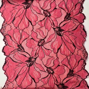 Op de afbeelding: Close-up van een bloemenkanten stof. Het kant heeft een herhalend patroon van grote, gestileerde bloemen in roze en zwarte tinten. De randen van de stof zijn geschulpt. De stof wordt waarschijnlijk gebruikt voor kleding of knutselprojecten.