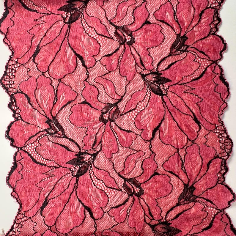 Op de afbeelding: Close-up van een bloemenkanten stof. Het kant heeft een herhalend patroon van grote, gestileerde bloemen in roze en zwarte tinten. De randen van de stof zijn geschulpt. De stof wordt waarschijnlijk gebruikt voor kleding of knutselprojecten.