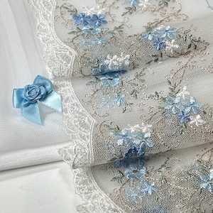 Könnte beinhalten: Nahaufnahme eines zarten weißen Spitzengewebes mit gestickten blauen Blumenmustern und einem gewellten Rand. Eine hellblaue Satinschleife mit Rosendetail ist am Stoff befestigt. Der Stoff ist gefaltet und zeigt Schichten.
