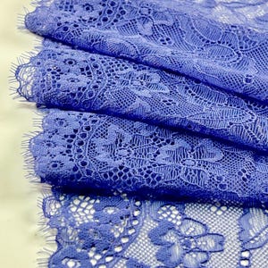 May include: Close-up of folded blue lace fabric. The lace features an intricate floral pattern with a scalloped edge. The fabric is a vibrant shade of blue, with visible texture and detail.
