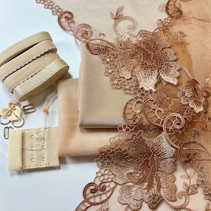 Könnte beinhalten: Eine Auswahl an beigen Dessous-Materialien. Enthält Rollen mit Gummiband, floraler Spitzenborte, Stoff, Haken- und Ösenverschlüssen und Metallbeschlägen. Die Spitze hat ein detailliertes Blumendesign in einem warmen Braunton. Der Stoff und das Gummiband sind in passenden neutralen Tönen gehalten.