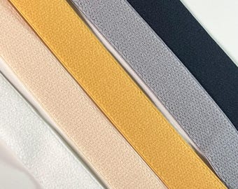 May include: Close-up of several elastic bands in various colors, including white, beige, gold, gray, and navy. The bands have a textured surface and are arranged diagonally across the frame, showcasing a range of hues and textures.