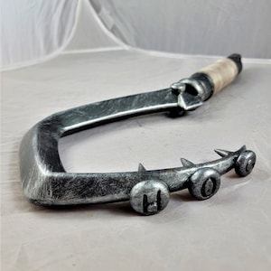 May include: A silver-coloured metal hook with a wooden handle. The hook has a series of spikes along the edge and three round metal accents with the letters "H.O.C." embossed on them.