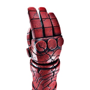 May include: A red, stone-like hand sculpture with a textured surface. The hand is in a fist position with the index finger pointing up. The sculpture is labeled "HEILBOI" at the base.