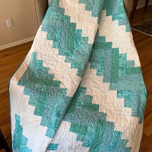 May include: A quilt featuring a log cabin block pattern in shades of teal, turquoise, and white. The design consists of rectangular fabric strips arranged in geometric steps, with intricate swirling machine quilting stitches across the surface.