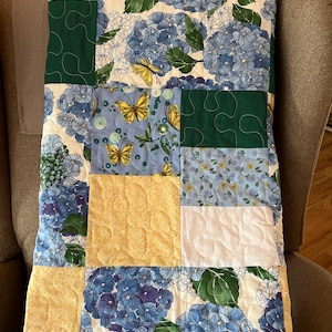 May include: A patchwork quilt featuring a floral pattern of blue hydrangeas and yellow butterflies on a white background. The quilt also includes green and yellow square patches. The quilt is draped over a gray armchair.