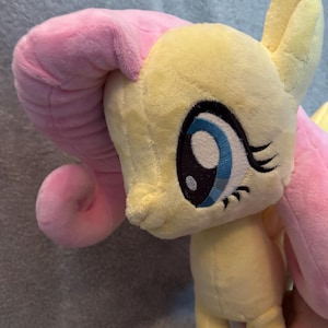 May include: A plush toy of a pony with a yellow body and long, soft pink hair. The pony features large, embroidered eyes with blue irises, black pupils, and long black eyelashes. The character has a small, pointed ear and a smooth, textured fabric finish.