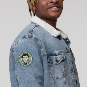 May include: A light blue denim jacket with a sherpa collar and a green embroidered patch featuring a cartoon character with black hair and a white shirt.