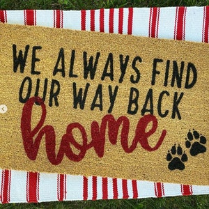 Op de afbeelding: Een rechthoekige deurmat met de tekst "WE ALWAYS FIND OUR WAY BACK HOME" in zwart en rood. Twee zwarte pootafdrukken staan rechts. De mat ligt op een rood-wit gestreept tapijt.