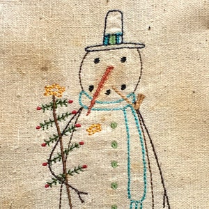 May include: Embroidered snowman with a blue scarf and a green and white hat holding a small green tree with red berries. The snowman is stitched on a beige fabric.
