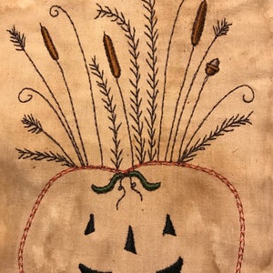 May include: Embroidered pumpkin with a jack-o'-lantern face, brown cattails, and other foliage. The pumpkin is outlined in red thread, with a green stem. The background is a light tan color.
