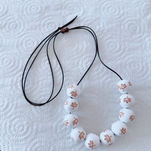 May include: A white beaded necklace with a black cord. Each white bead is decorated with a brown maple leaf design. The necklace is displayed on a white, textured surface.