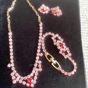 Könnte beinhalten: Ein Vintage-Schmuckset mit Halskette, Armband und Ohrringen. Das Set hat eine goldfarbene Metalleinfassung mit rosa und roten Strasssteinen. Die Halskette und das Armband haben ein zartes Design, und die Ohrringe sind klein und rund.