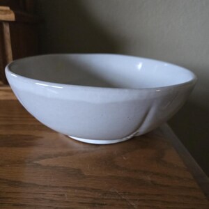May include: A white ceramic bowl with a rounded shape. The bowl has a smooth, glossy finish and sits on a wooden surface. The bowl is empty and appears to be clean.