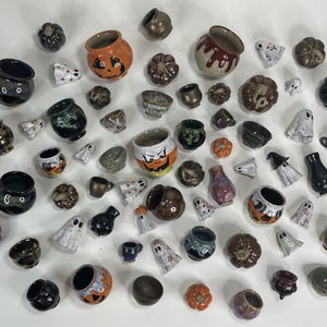 May include: A collection of miniature ceramic items including small cauldrons, pumpkins, and ghost figures. The pieces feature various glazes in black, orange, brown, and white, with some items decorated with painted jack-o'-lantern faces.