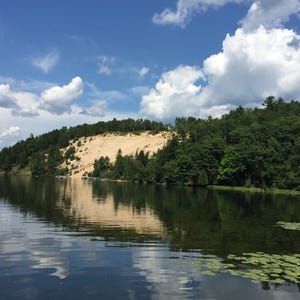 May include: A sandy cliff with lush green trees on top, overlooking a calm lake with a blue sky and white clouds. The lake is reflecting the sky and clouds.