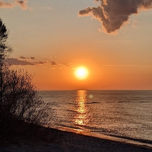 Può includere: Un tramonto vibrante su uno specchio d'acqua calmo con una spiaggia sabbiosa in primo piano. Il sole tramonta dietro una grande nuvola, proiettando un bagliore caldo sulla scena.
