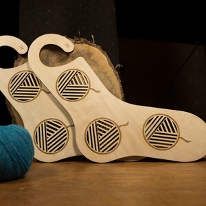 May include: Two wooden sock holders with a yarn ball design. The holders are light brown and have a hook at the top. The holders are sitting on a wooden surface with two balls of yarn.