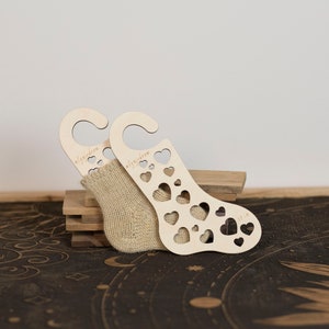 May include: A pair of wooden sock blockers with heart cutouts and a beige sock. The sock blockers have a curved top with a hole for hanging. The sock is partially inserted into one of the blockers. The text "algidream" is visible on the sock blockers.