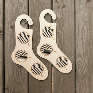 May include: Two wooden sock-shaped holders with a hook at the top. Each holder has a cutout design of balls of yarn.