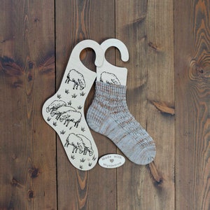 May include: Two wooden sock hangers featuring a sheep design. One hanger has a pair of knitted socks hanging on it. The socks are a light brown and white colour with a textured pattern. The hangers have a small heart with the text "Made with Mlyniedrow" on them.