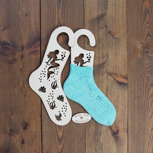 Può includere: Due portacalze in legno bianco a forma di calze con un'immagine di sirena ritagliata. I portacalze sono su una superficie di legno marrone. Un portacalze ha un calzino di lana blu sopra. Il calzino è di colore blu chiaro con un motivo a pizzo. Il portacalze ha il testo "Made with love Mlynjadrow".